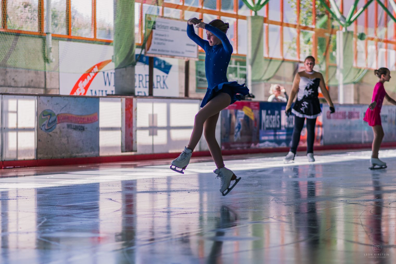 Eiskunstläuferin im Sprung in der Eissporthalle, dahinter weitere Eiskunstläufer.