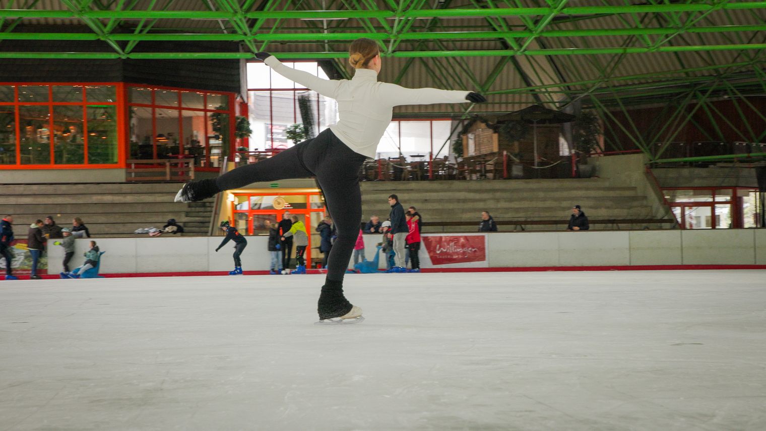 Eiskunstläuferin hebt zur Drehung an, im Hintergrund mehrere Gäste auf der Eisfläche.