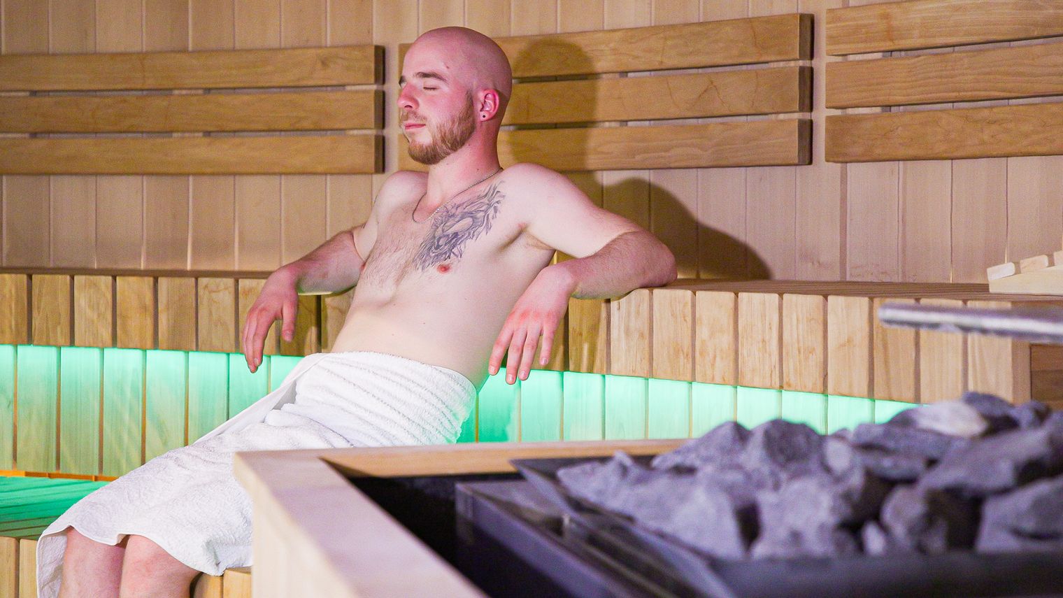 Ein Mann im Badetuch bekleidet sauniert in der Finnischen Sauna, im Vordergrund sieht man den Saunaofen.