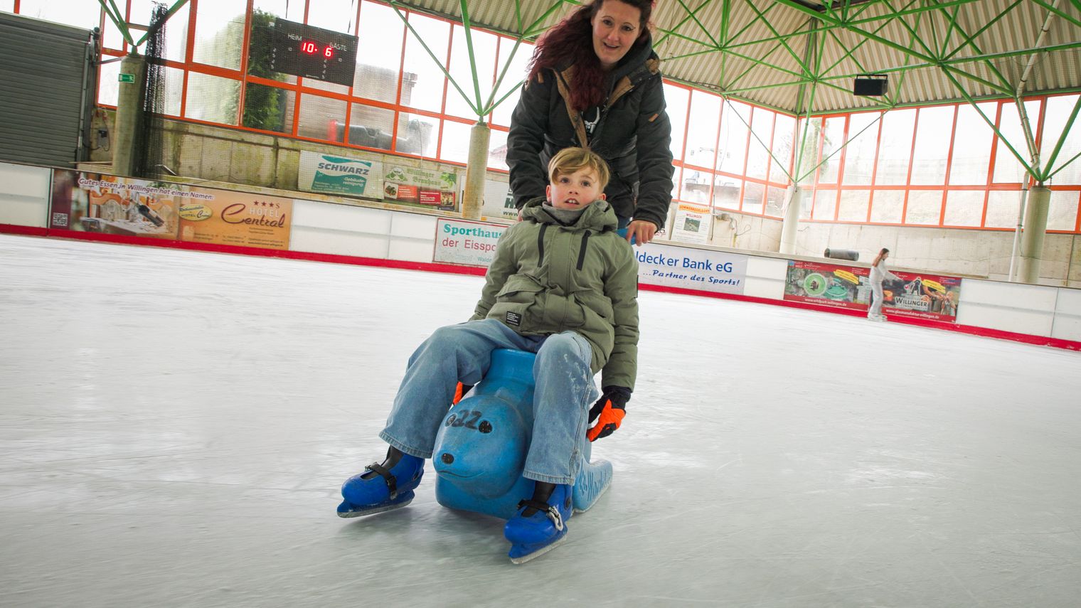Mutter schiebt ihr Kind, dass auf der Lauflernhilfe in Form einer Robbe sitzt, über die Eisfläche.