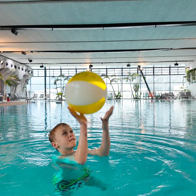 Kind spielt mit einem weiß-gelben Wasserball im Solebecken, im Hintergrund sind Liegen und Palmen zu sehen.
